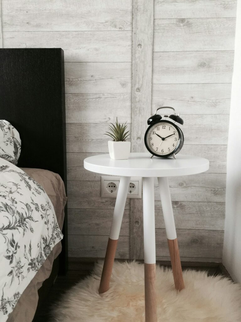 Uma mesa de cabeceira ao lado da cama. Ela tem o tampo redondo e está com uma pequeno vaso de planta comportando uma leguminosa e uma relógio antigo.