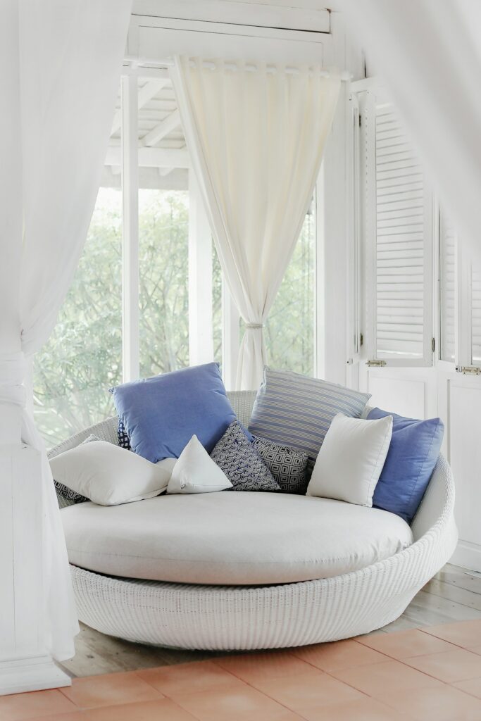 Uma linda poltrona cinza com almofadas azuis e brancas. Ao fundo uma janela com jardim e uma bela cortina branca